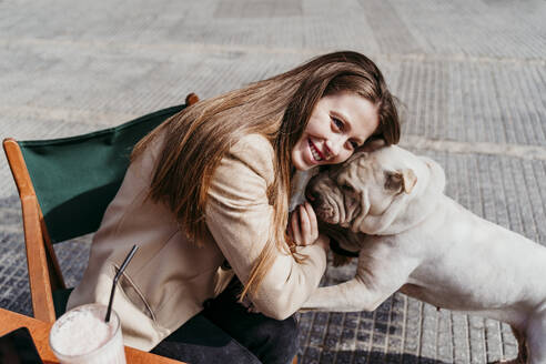 Lächelnde Frau lehnt sich an ein Haustier, während sie in einem Straßencafé sitzt - EBBF03487