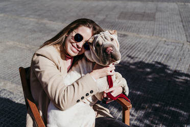 Smiling young woman embracing dog while sitting on chair - EBBF03493