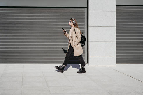 Junge Frau mit Kopfhörern und Mobiltelefon auf dem Gehweg - EBBF03497