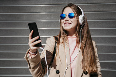 Schöne Frau mit Sonnenbrille und Kopfhörern, die ein Smartphone an einem sonnigen Tag benutzt - EBBF03498