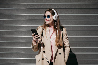 Young woman with headphones and mobile phone standing in front of shutter - EBBF03499