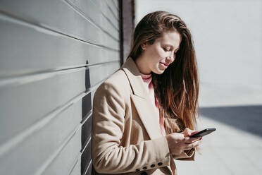 Young woman using mobile phone by shutter on sunny day - EBBF03502