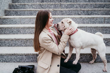 Smiling woman looking at pet while sitting on steps - EBBF03503