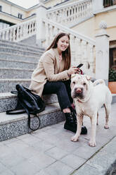 Smiling woman with mobile phone sitting by purebred dog on steps - EBBF03504