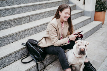 Beautiful woman with Shar-Pei dog using mobile phone while sitting on staircase - EBBF03507