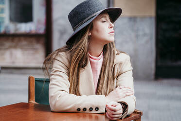 Young woman wearing hat sitting with eyes closed at sidewalk cafe - EBBF03518