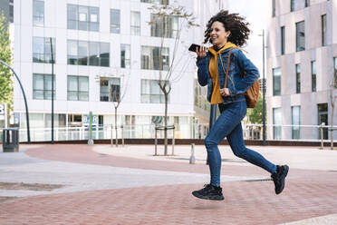 Lächelnde junge Frau mit Rucksack, die beim Laufen in der Stadt mit ihrem Handy telefoniert - JCZF00702