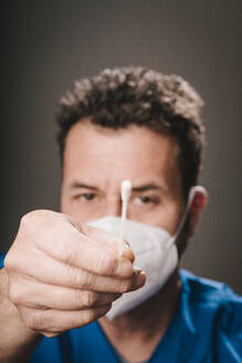 Männlicher Mitarbeiter im Gesundheitswesen mit Gesichtsschutzmaske, der einen Nasenabstrich vor einem grauen Hintergrund zeigt - MMFF01302