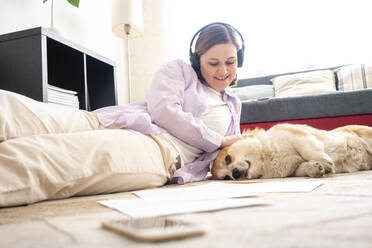 Smiling woman wearing headphones stroking dog while relaxing on carpet at home - VPIF04006