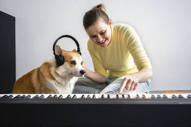 Lächelnde Frau mit Hund, die dem Hund Kopfhörer aufsetzt, während sie im Wohnzimmer Klavier übt - VPIF04033