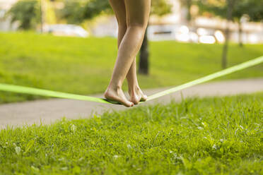 Junge Frau balanciert barfuß auf einer Slackline in einem öffentlichen Park - MTBF01011