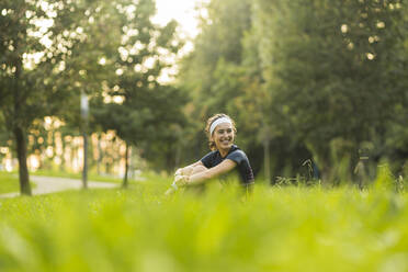 Lächelnde Frau, die ihre Knie umarmt, während sie in einem öffentlichen Park auf dem Rasen sitzt - MTBF01015