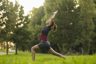 Young woman practicing warrior pose yoga at public park - MTBF01016