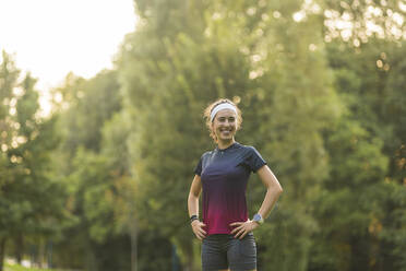 Smiling woman with hands on hip standing in public park - MTBF01021