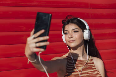 Lächelndes Teenager-Mädchen, das ein Selfie über ein Smartphone macht, während es Musik hört - JRVF00591