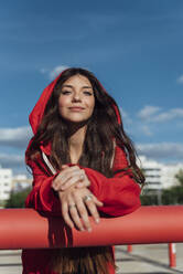 Lächelnder weiblicher Teenager in roter Jacke, der sich an ein Rohr lehnt, an einem sonnigen Tag - JRVF00595