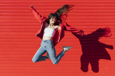 Cheerful teenage girl jumping in front of red wall - JRVF00598