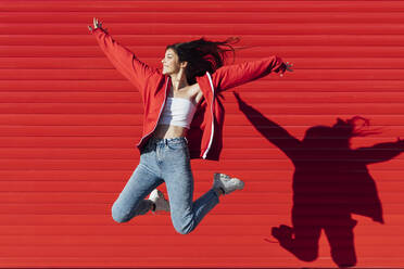 Smiling teenage girl with arms outstretched jumping in front of red wall - JRVF00599
