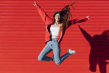 Teenage girl with tousled hair jumping in front of red wall - JRVF00600