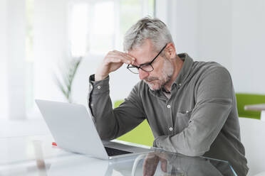 Overworked businessman with head in hand sitting in front of laptop at desk - MCVF00846