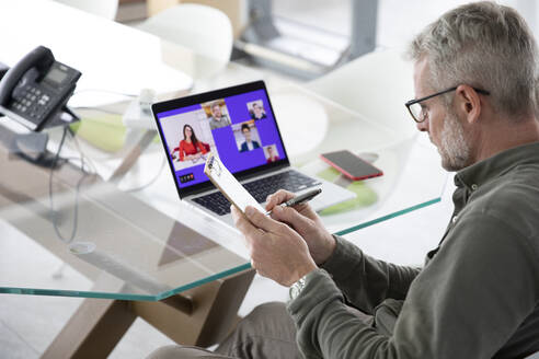 Männlicher Fachmann, der während einer Videokonferenz über einen Laptop im Büro auf ein Klemmbrett schreibt - MCVF00848