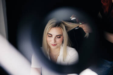 Female hairstylist using straightener on beautiful model's hair seen through ring light - MRRF01124