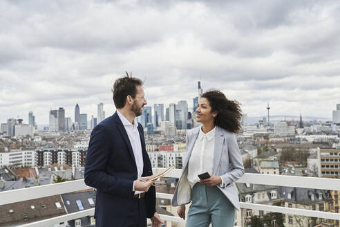 Male and female partners discussing while standing on rooftop - AKLF00191