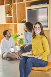 Geschäftsfrau lächelt, während sie mit Kollegen im Hintergrund in einem kreativen Büro sitzt - AKLF00208