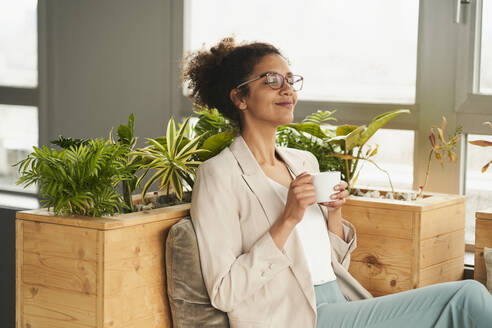 Smiling woman with coffee cup sitting in office - AKLF00235