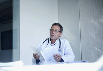 Thoughtful male doctor with medical report contemplating while sitting in hospital - AJOF01308