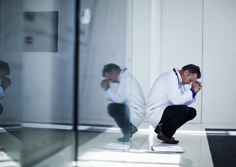 Stressed male doctor with head in hands crouching by medical reports in hospital - AJOF01324