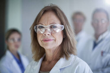 Female doctor wearing protective glasses with colleagues in background at hospital - AJOF01338