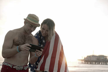 Gut aussehender Mann, der am Strand von Santa Monica mit einer in eine Flagge gehüllten Freundin ein digitales Tablet benutzt - AJOF01358