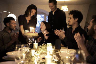 Woman blowing candles on birthday cake amidst male and female friends clapping during celebration at home - MJRF00443