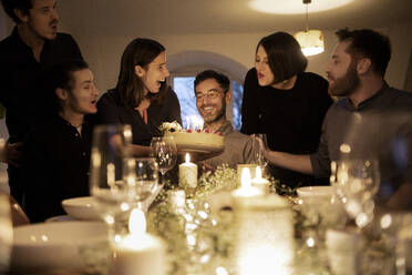 Male and female friends cheering during birthday celebration of smiling man at home - MJRF00445