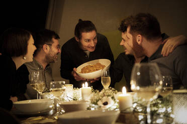 Smiling man offering pasta to male and female friends at home - MJRF00453