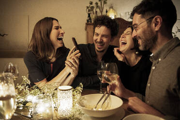 Männliche und weibliche Freunde lachen, während die Frau am Esstisch ihr Handy zeigt - MJRF00459