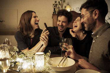 Male and female friends laughing while woman showing mobile phone at dining table - MJRF00459