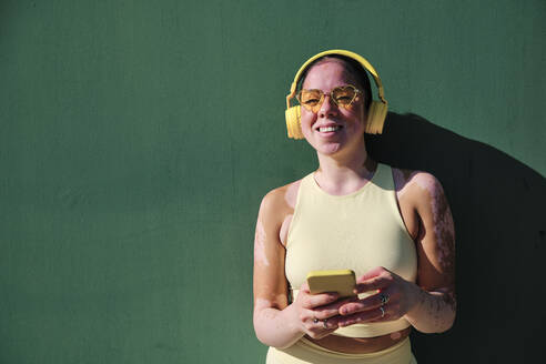 Smiling woman with eyeglasses holding smart phone by green wall - AGOF00104