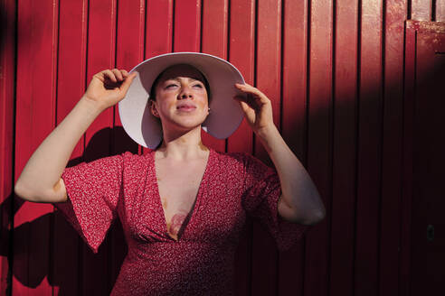 Young woman looking away wearing sun hat by red wall - AGOF00109