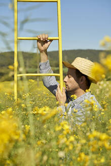 Serious man with straw hat holding ladder in field during sunny day - VEGF04491