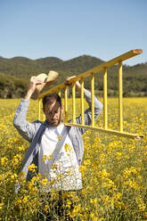Junger Mann, der eine Leiter trägt, während er in einem gelben Feld spazieren geht - VEGF04504