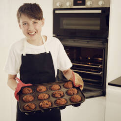Lächelnder Junge hält ein Tablett mit frisch gebackenen Muffins in der Küche zu Hause - PWF00322