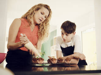 Sohn schaut Mutter an, die Muffins mit einem Spritzbeutel in der Küche glasiert - PWF00324