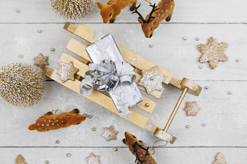 Weihnachtsgeschenk auf Spielzeug-Schlitten, umgeben von Spielzeug-Hirschen mit selbstgebackenen Keksen vor weißem Holzhintergrund - GWF07022
