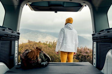 Back view of male camper in outerwear standing near van and admiring scenic view of highlands - ADSF24517
