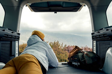 Back view of unrecognizable male camper in outerwear lying down on van and admiring scenic view of highlands - ADSF24520