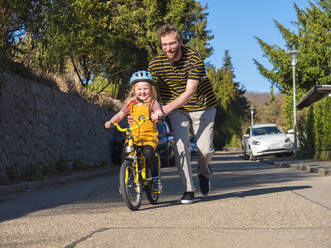 Lächelnder Vater, der seine Tochter beim Radfahren auf der Straße unterstützt - LAF02714