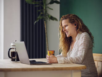 Smiling woman having coffee while looking at laptop - PWF00332