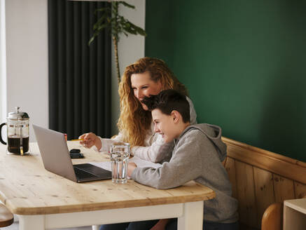 Lächelnde Frau, die ihrem Sohn beim E-Learning am Laptop zu Hause hilft - PWF00333